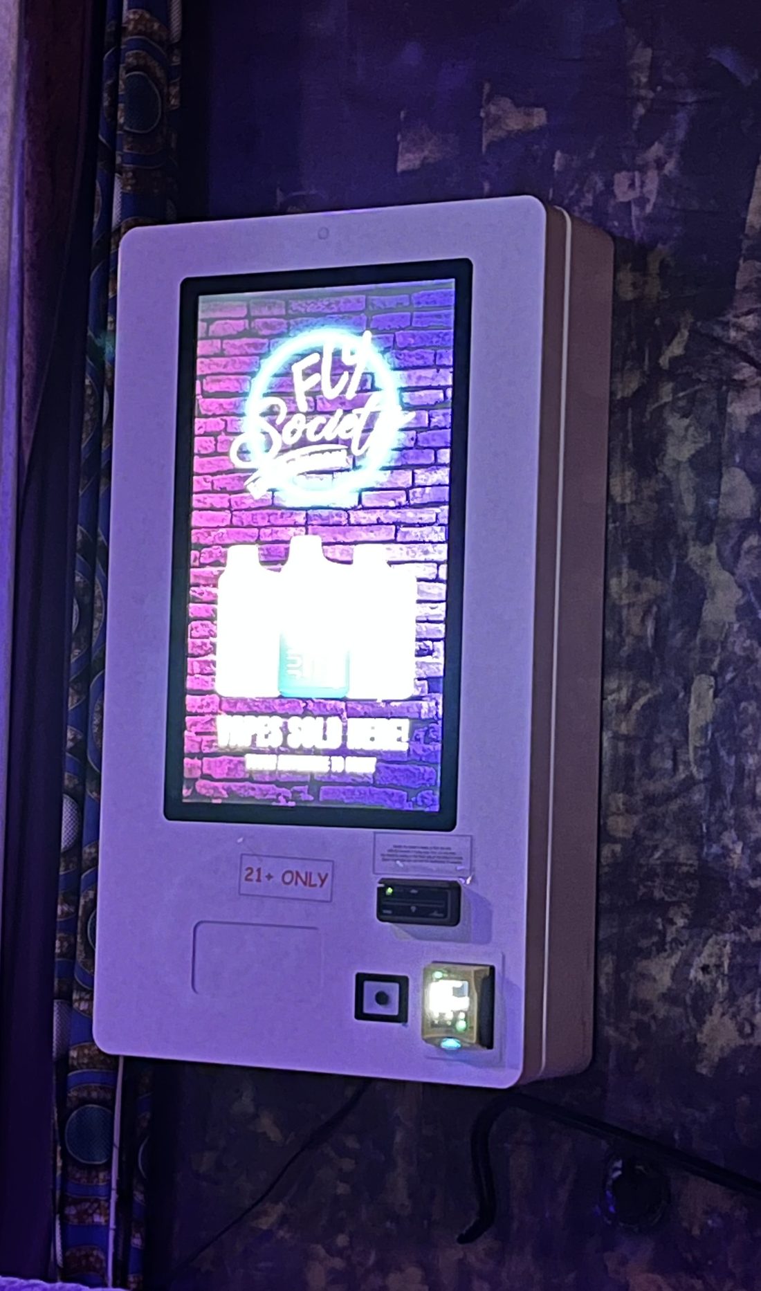 Fly Society vape vending machine mounted in a nightclub under purple lighting, with a 21+ only label